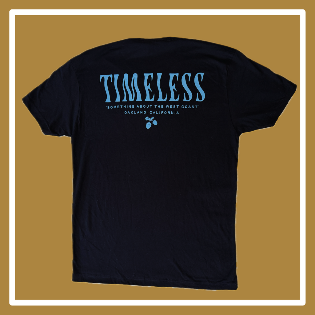 A black t-shirt laying on a golden and white background, the front has a blue scorpion in the upper right hand corner (of the photo, left hand corner on the shirt itself); the back reads “Timeless” in a squiggly blue font, and “something about the west coast, Oakland, California,” underneath this all is small blue acorns.