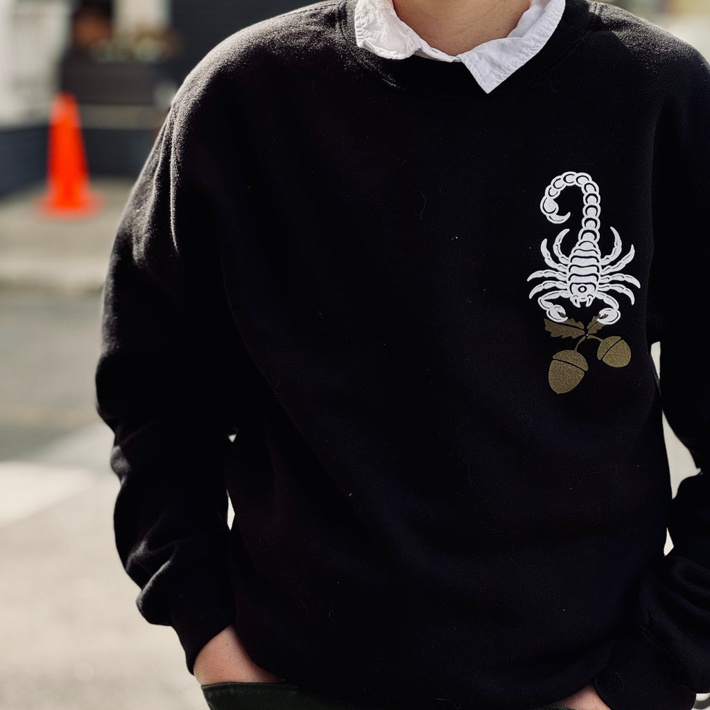 A person wears a white collared shirt underneath a black crewneck sweatshirt; the front has a white scorpion holding golden acorns in the upper right hand corner (of the photo, left hand corner on the shirt itself). Their hands are in both jeans pockets.