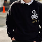 A person wears a white collared shirt underneath a black crewneck sweatshirt; the front has a white scorpion holding golden acorns in the upper right hand corner (of the photo, left hand corner on the shirt itself). Their hands are in both jeans pockets.