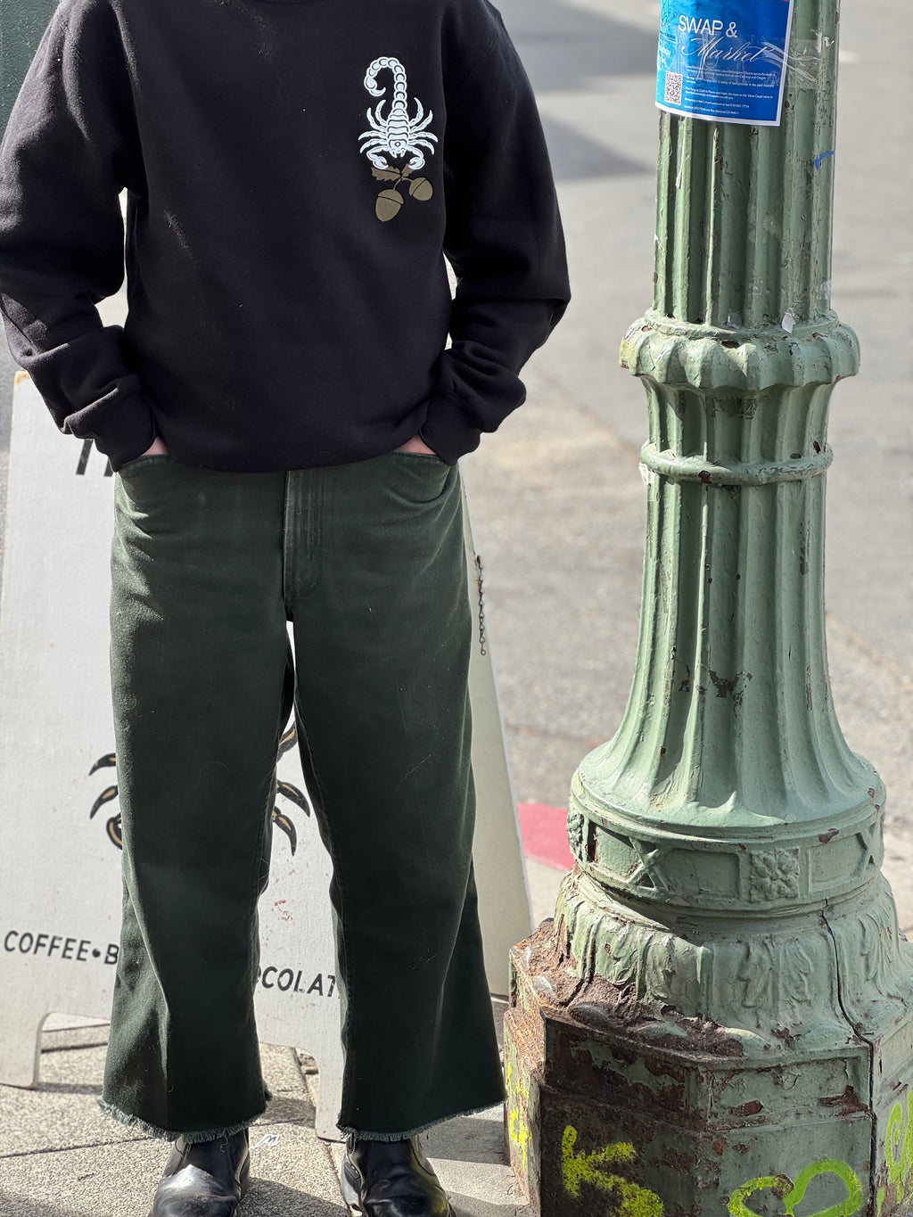 A person stands next to a green lamppost, wearing green jeans, and a black crewneck sweatshirt; the front has a white scorpion holding golden acorns in the upper right hand corner (of the photo, left hand corner on the shirt itself). Their hands are in both jeans pockets.