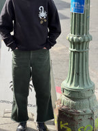 A person stands next to a green lamppost, wearing green jeans, and a black crewneck sweatshirt; the front has a white scorpion holding golden acorns in the upper right hand corner (of the photo, left hand corner on the shirt itself). Their hands are in both jeans pockets.