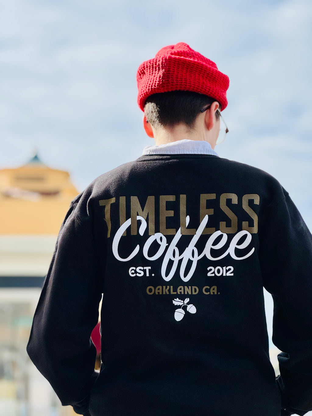 A person facing away with a red beanie and glasses, is wearing a black crewneck sweatshirt; the back reads “Timeless Coffee” in a gold and white font, and “est. 2012, Oakland, CA,” underneath this all is small white acorns. 