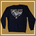A black crewneck sweatshirt laying on a golden and white background, the front has a white scorpion holding golden acorns in the upper right hand corner (of the photo, left hand corner on the shirt itself); the back reads “Timeless Coffee” in a gold and white font, and “est. 2012, Oakland, CA,” underneath this all is small white acorns. 