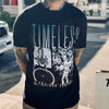 A person, heavily tattooed, standing with their arms behind them, and wearing a black t-shirt with the front reading “Timeless, Oakland Forever” and images of a scorpion, an Oakland A’s baseball player, and a bicycle wheel.