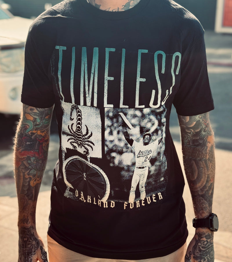 A person, heavily tattooed, standing with their arms to the side, and wearing a watch; wearing a black t-shirt with the front reading “Timeless, Oakland Forever” and images of a scorpion, an Oakland A’s baseball player, and a bicycle wheel.