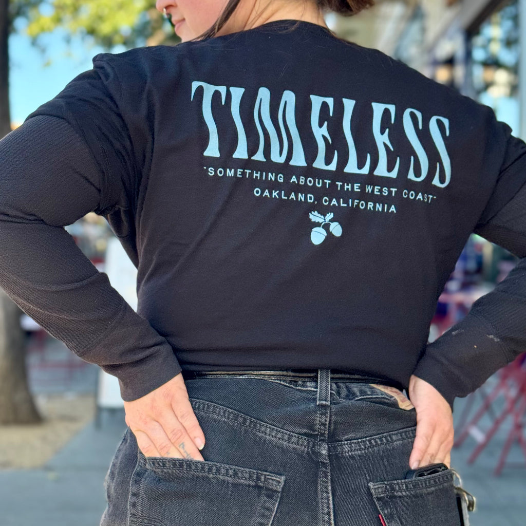A person, facing away, with both hands in their jeans pockets, wears a black t-shirt with the back reading “Timeless” in a squiggly blue font, and “something about the west coast, Oakland, California,” underneath this all is small blue acorns. The shirt is cropped by tying it at the waist - just above a belt holding up their jeans.