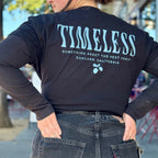 A person, facing away, with both hands in their jeans pockets, wears a black t-shirt with the back reading “Timeless” in a squiggly blue font, and “something about the west coast, Oakland, California,” underneath this all is small blue acorns. The shirt is cropped by tying it at the waist - just above a belt holding up their jeans.