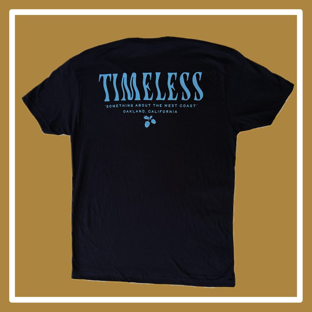 A black t-shirt laying on a golden and white background, the front has a blue scorpion in the upper right hand corner (of the photo, left hand corner on the shirt itself); the back reads “Timeless” in a squiggly blue font, and “something about the west coast, Oakland, California,” underneath this all is small blue acorns. 