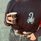A person with one hand in their jeans pocket, and one hand holding a black mug, also wearing a black t-shirt with the front having a blue scorpion in the upper right hand corner (of the photo, left hand corner on the shirt itself); the shirt is cropped by tying it at the waist - just above a belt holding up their jeans.