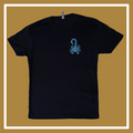 A black t-shirt laying on a golden and white background, the front has a blue scorpion in the upper right hand corner (of the photo, left hand corner on the shirt itself); the back reads “Timeless” in a squiggly blue font, and “something about the west coast, Oakland, California,” underneath this all is small blue acorns. 