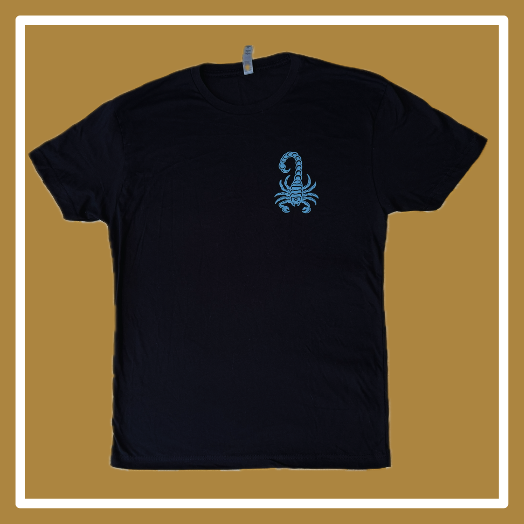 A black t-shirt laying on a golden and white background, the front has a blue scorpion in the upper right hand corner (of the photo, left hand corner on the shirt itself); the back reads “Timeless” in a squiggly blue font, and “something about the west coast, Oakland, California,” underneath this all is small blue acorns. 