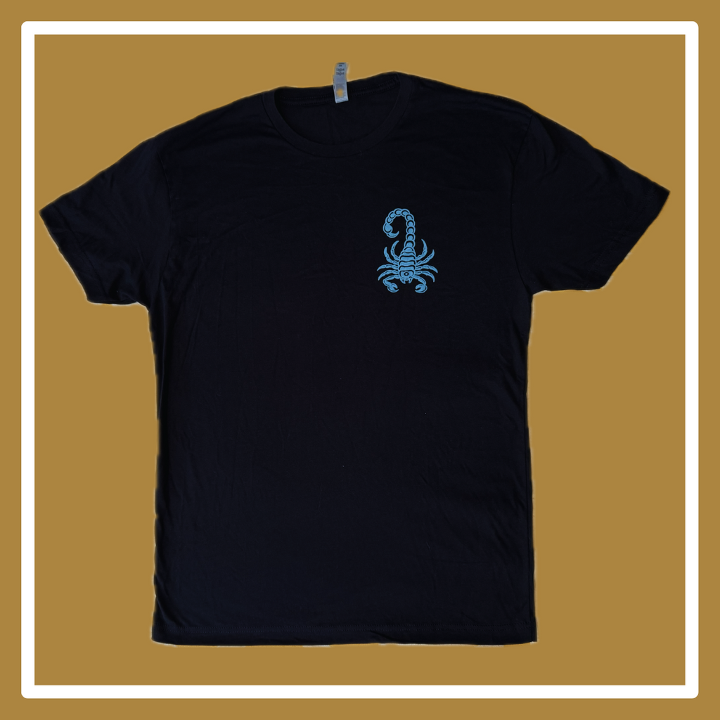 A black t-shirt laying on a golden and white background, the front has a blue scorpion in the upper right hand corner (of the photo, left hand corner on the shirt itself); the back reads “Timeless” in a squiggly blue font, and “something about the west coast, Oakland, California,” underneath this all is small blue acorns. 